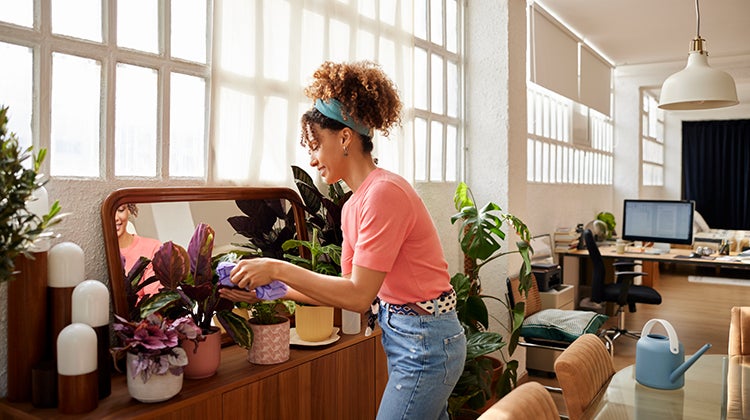 Woman happily watering her plants in her main living space as the sun shines in from the windows