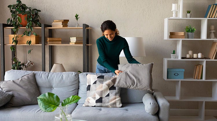 Clear out the clutter Woman standing behind a couch, organizing the the pillows that are laying on top of it.