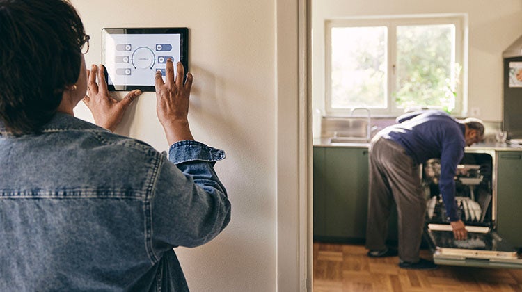 Make your home energy efficient In the first plane we see a woman from behind, she is adjusting a thermostat that is attached to a white wall. We see an older man in the next room, he is loading a dishwasher