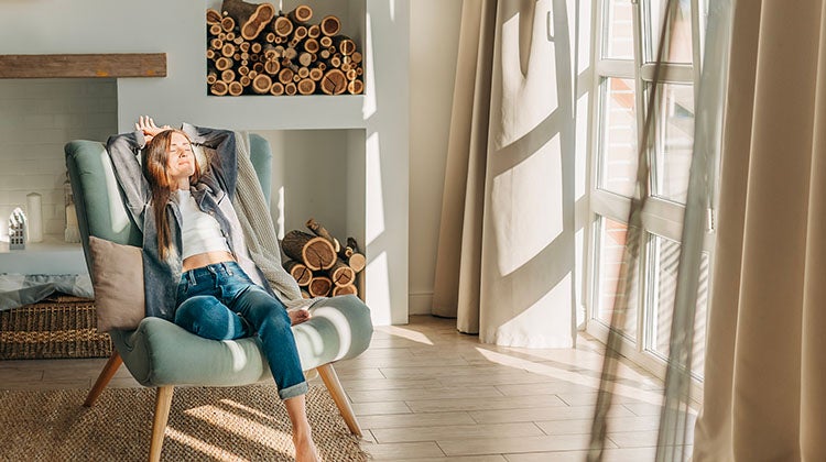 Embrace elements of minimalism woman siting in an armchair in front of a fireplace, relaxing and enjoying the sunset lights coming in through a big window beside her. The room looks very clean and organized.
