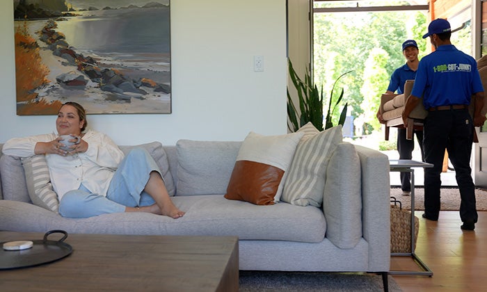 A woman relaxing on her couch while two 1-800-GOT-JUNK? Truck Team Members remove her junk.