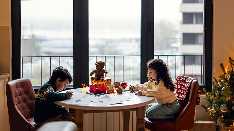 Two young children writing holiday letters by a large window